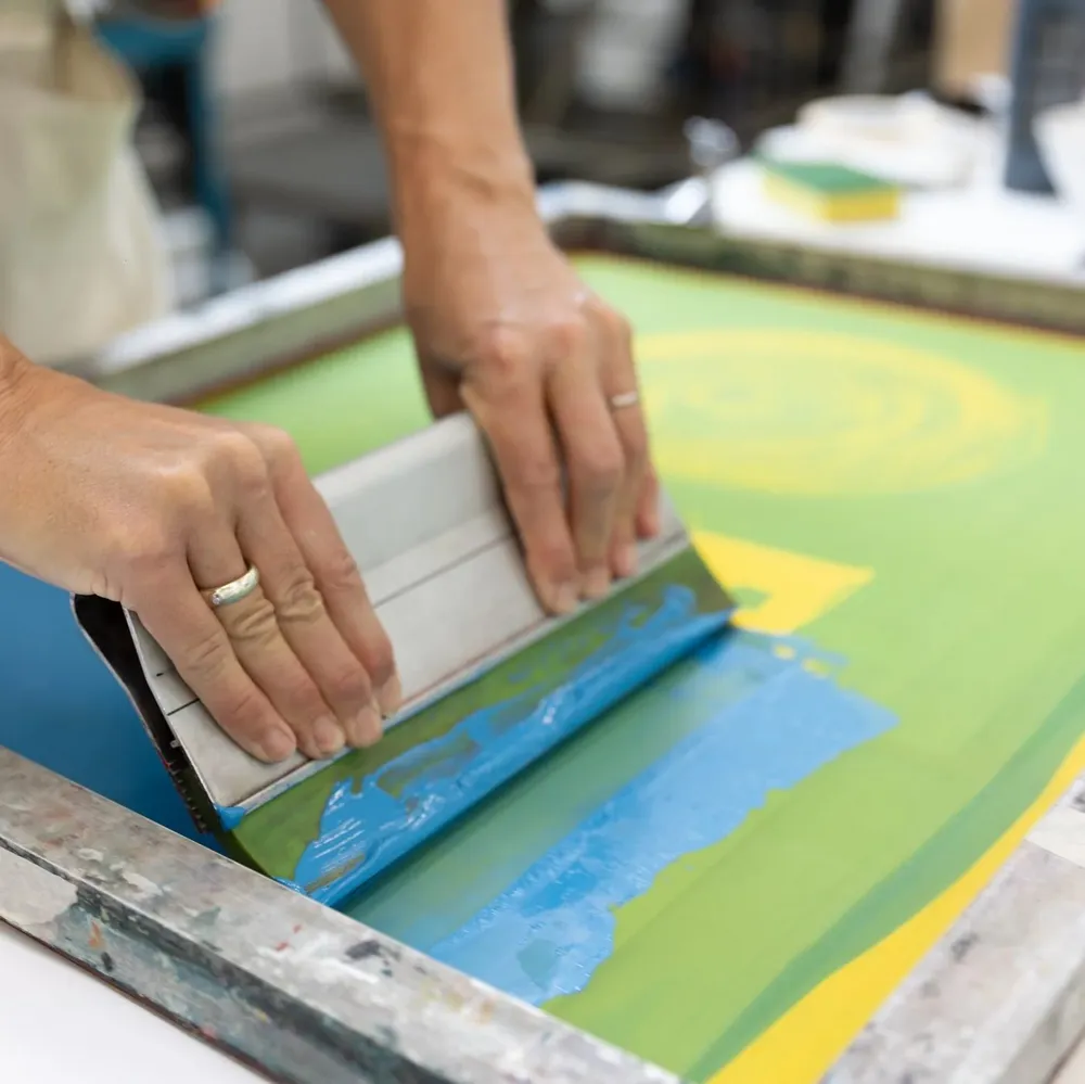 Hands pulling a squeegee through blue ink across a printing screen
