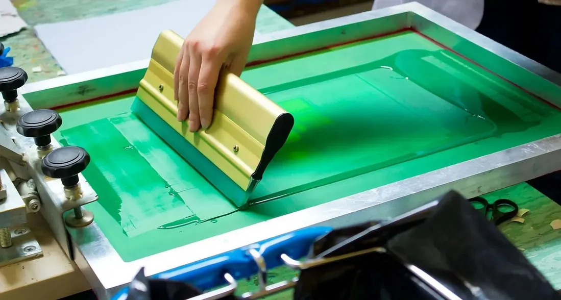 A hand pulling a squeegee through bright green ink on a screen printing frame